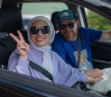 Mona and Sebatian holding hands while sitting in their car. Both smile at the camera, and Mona flashes a peace sign. Photo courtesy of Unity Productions Foundation and PBS